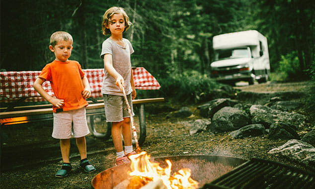 Two kids around a campfire with an RV in the background